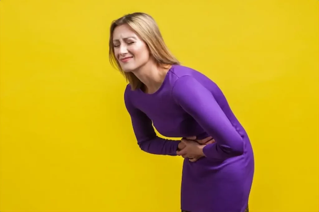 Portrait of unhealthy distressed woman in elegant purple dress clutching her belly, grimacing from acute abdominal pain, gastritis or constipation. indoor studio shot isolated on yellow background