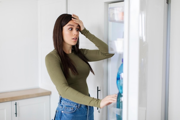 Shocked woman looking into fridge displaying surprise and disbelief about lack of food home kitchen