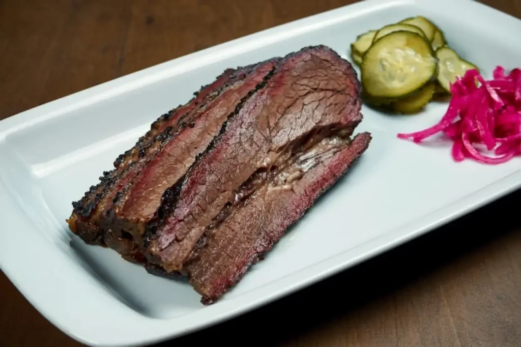 Close up view on tasty brisket beef from smoker on white plate on wooden surface. Grilled meat pastrami