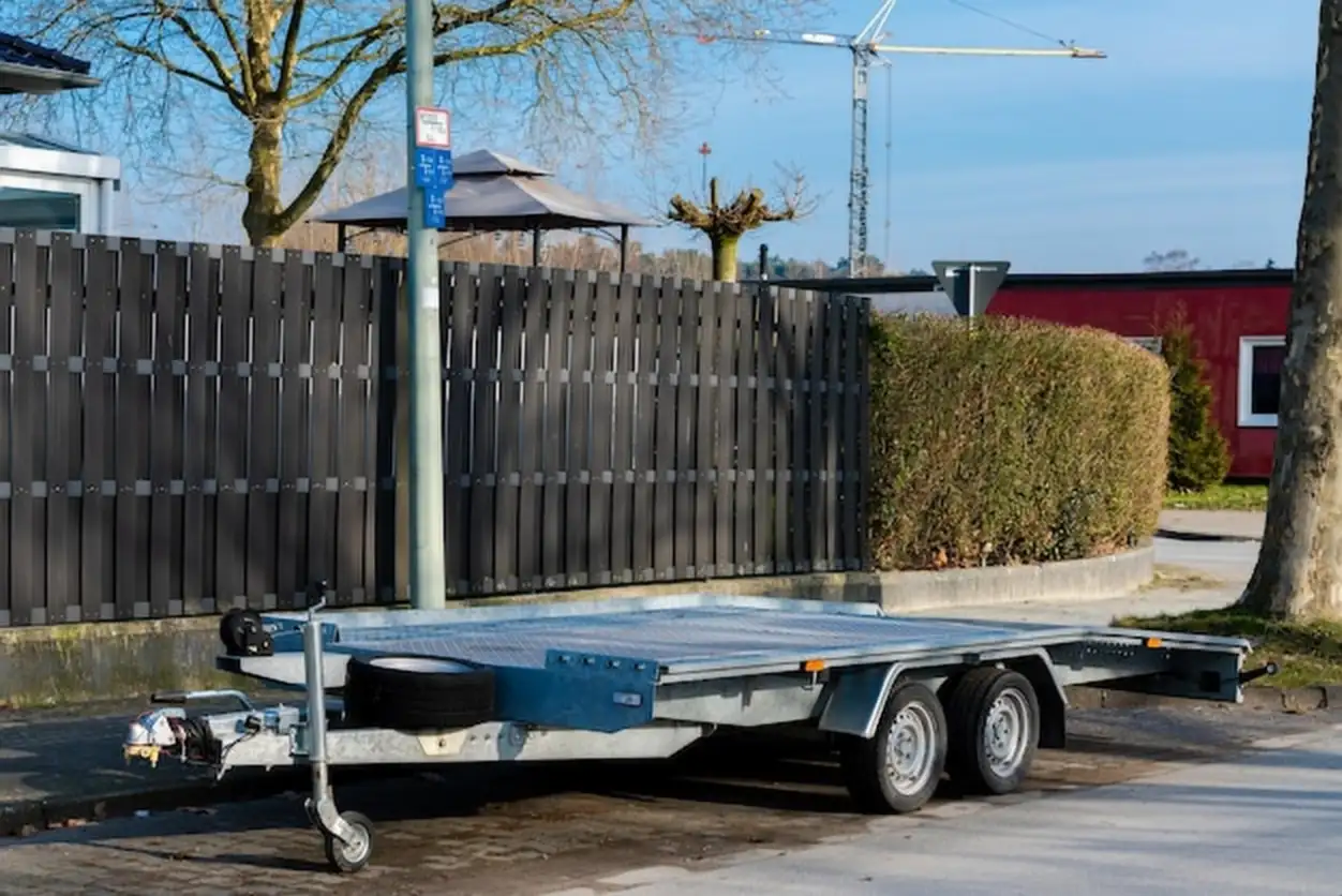 A metallic trailer is parked next to a hedge with construction cranes visible in the background the