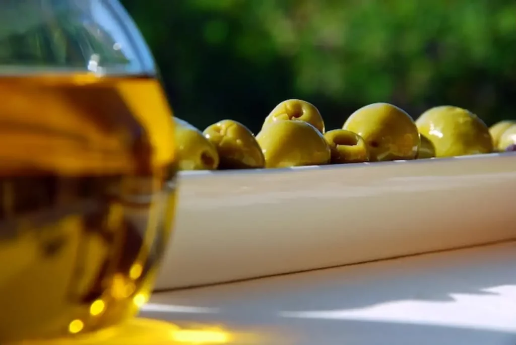 A bowl of olives with a glass of olive oil in the background.