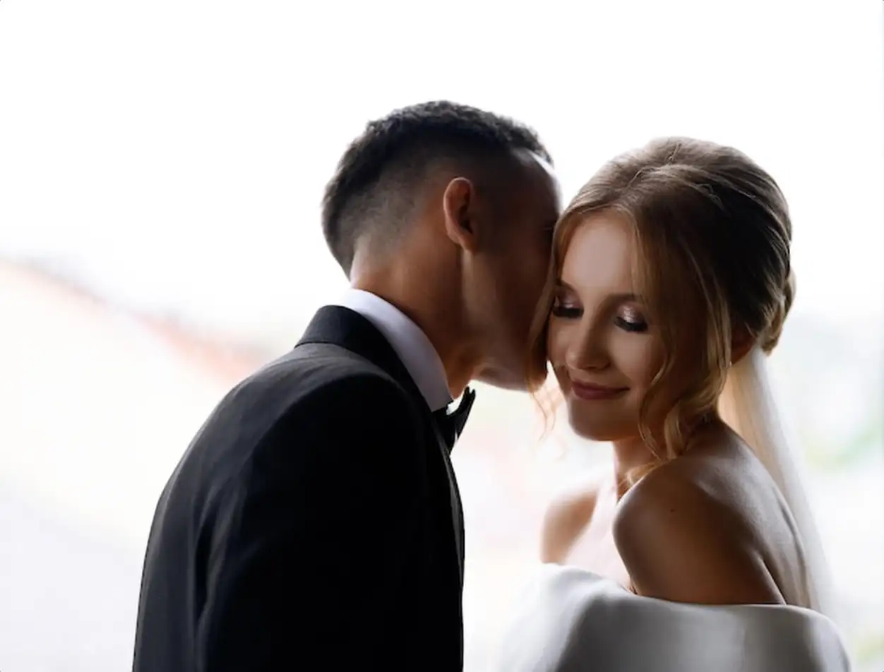 Close view of pretty woman with stylish hairdo dressed in white gown with naked shoulders smiling and looking away while faceless groom kissing her face during wed celebration