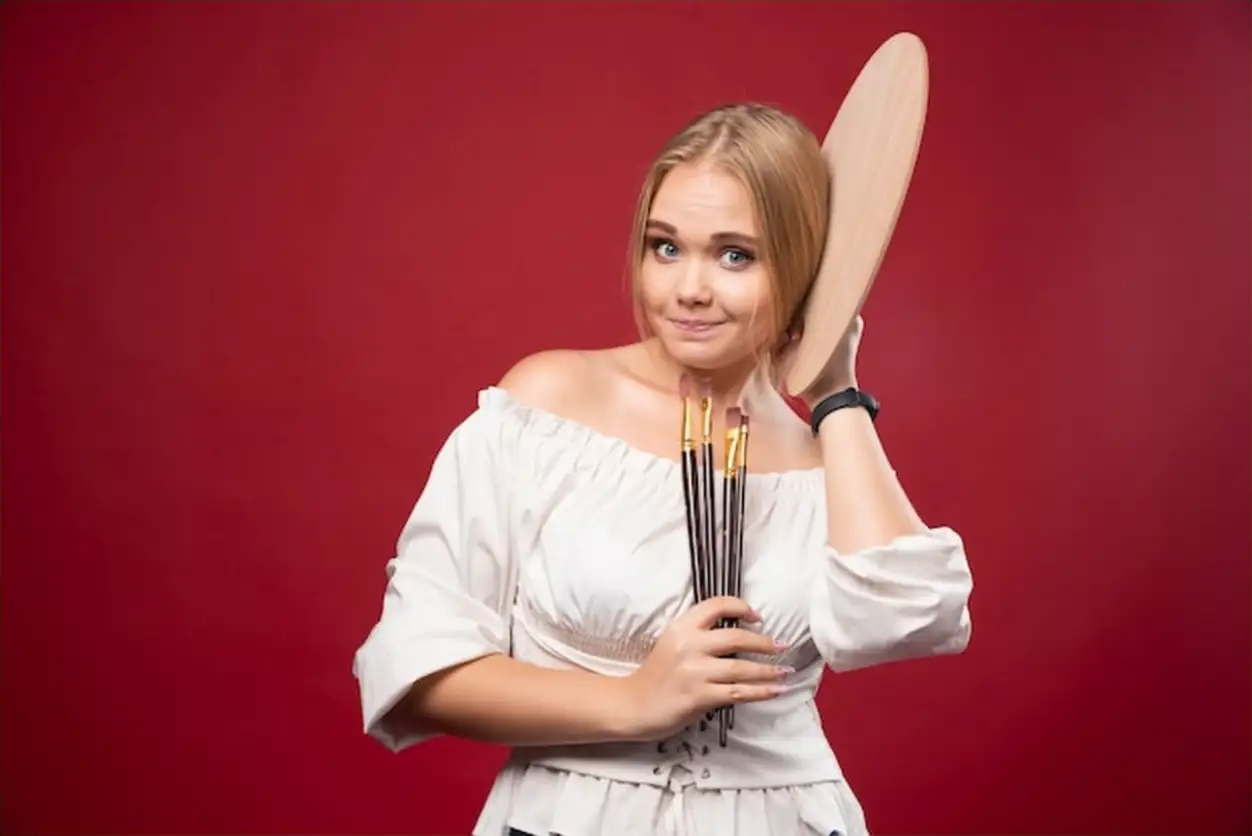 Blonde artist holds a palette and brushes and looks positive and dedicated.
