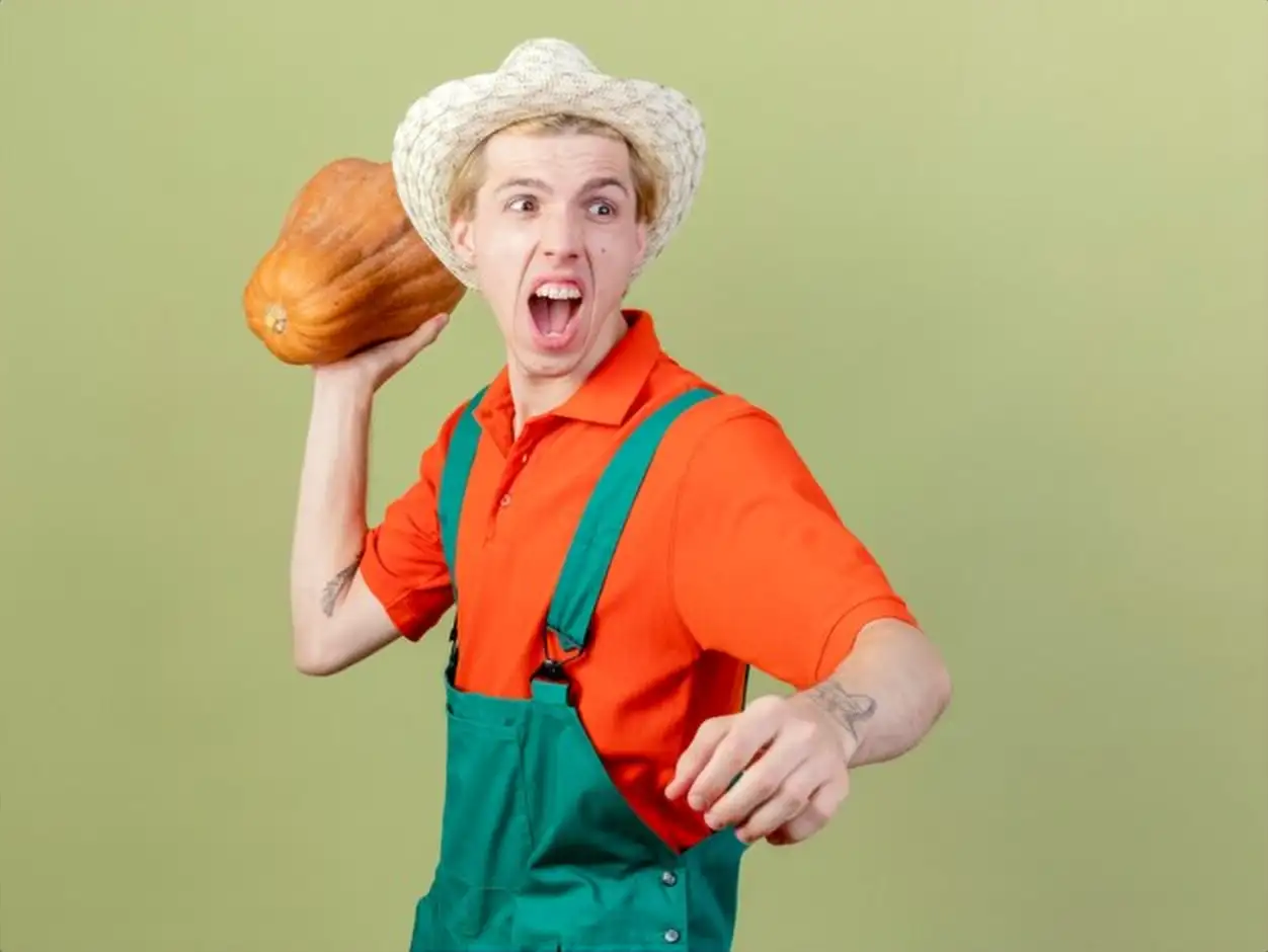 Young gardener man wearing jumpsuit and hat swinging pumpkin shouting with aggressive expression standing over light background