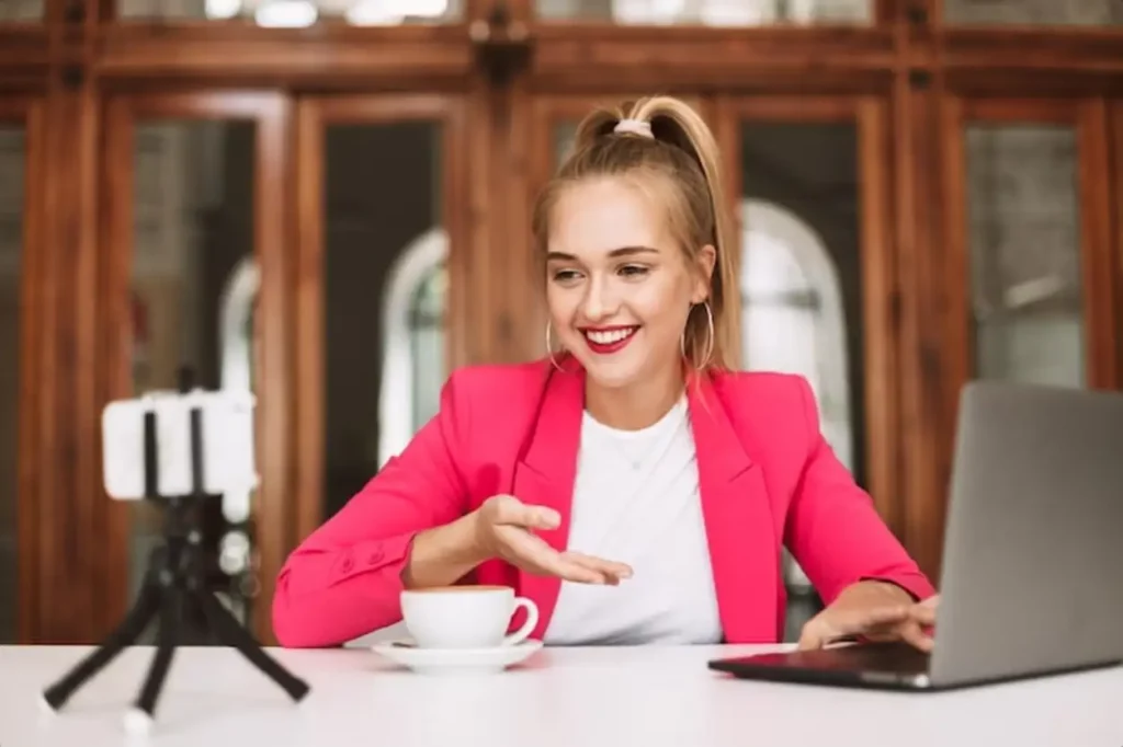 Happy girl in pink jacket with cup of coffee and laptop joyfully recording new video for her vlog
